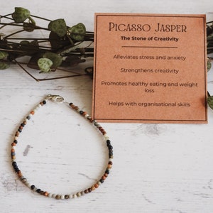 May include: A brown card with the text "Picasso Jasper, The Stone of Creativity" and a list of benefits: Alleviates stress and anxiety, Strengthens creativity, Promotes healthy eating and weight loss, Helps with organisational skills. A brown and white beaded bracelet with a silver clasp.