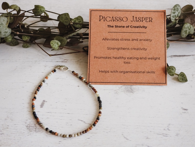 May include: A brown card with the text "Picasso Jasper, The Stone of Creativity" and a list of benefits: Alleviates stress and anxiety, Strengthens creativity, Promotes healthy eating and weight loss, Helps with organisational skills. A brown and white beaded bracelet with a silver clasp.