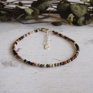 May include: A delicate anklet made with small, colorful beads in shades of brown, gray, black, and white. The anklet has a silver clasp.