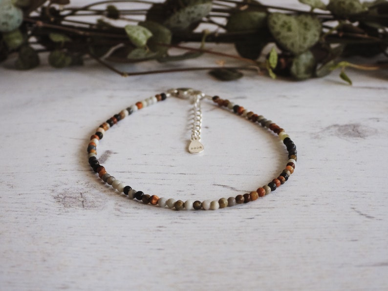 May include: A delicate anklet made with small, colorful beads in shades of brown, gray, black, and white. The anklet has a silver clasp.