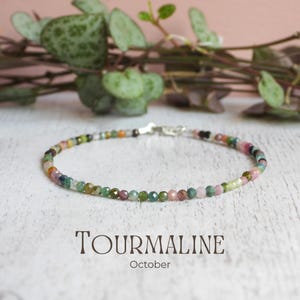 May include: A delicate bracelet composed of small, faceted, multi-coloured tourmaline beads in shades of pink, green, and brown. The bracelet features a silver-toned metal clasp and is labelled with the text TOURMALINE October.
