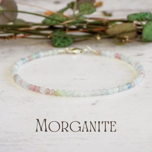 Op de afbeelding: Een delicate armband gemaakt van kleine, ronde, pastelkleurige morganiet kralen. De armband ligt op een wit houten oppervlak met het woord "MORGANITE" eronder.