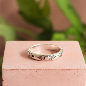 May include: A silver ring with an engraved pattern of sunburst and leaf-like designs. The ring is displayed on a pink, rectangular block, with a blurred green plant in the background. The ring is a band style.