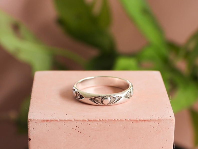 May include: A silver ring with an engraved pattern of sunburst and leaf-like designs. The ring is displayed on a pink, rectangular block, with a blurred green plant in the background. The ring is a band style.