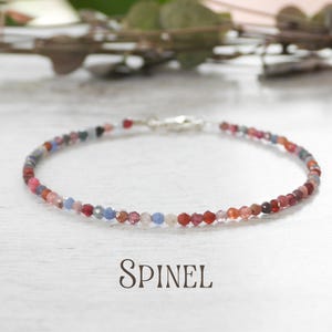 Könnte beinhalten: Ein zartes Armband aus kleinen, mehrfarbigen Spinellperlen. Die Perlen sind in Rot-, Rosa-, Blau- und Grautönen gehalten und mit einem silbernen Verschluss aufgefädelt. Das Wort "SPINEL" ist darunter gedruckt.