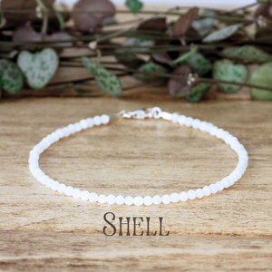 May include: A white shell bead anklet with a silver clasp. The anklet is on a wooden surface with green leaves in the background. "SHELL" is written in cursive below the anklet.