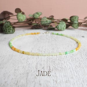 May include: A delicate jade necklace with a silver clasp. The necklace features small, colourful beads in shades of green, yellow, and orange. The word "JADE" is printed in white below the necklace.