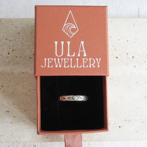 May include: A silver ring with an etched design is displayed inside an open, rectangular, coral-colored box. The box has the words "ULA JEWELLERY" printed in white. The ring is centered in the black interior of the box. The box is on a light-colored, textured surface.