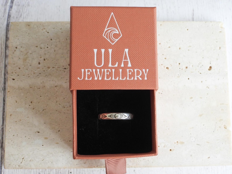 May include: A silver ring with an etched design is displayed inside an open, rectangular, coral-colored box. The box has the words "ULA JEWELLERY" printed in white. The ring is centered in the black interior of the box. The box is on a light-colored, textured surface.