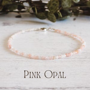 Könnte beinhalten: Eine zarte Halskette aus rosa Opal mit einem silbernen Verschluss. Die Halskette besteht aus kleinen, runden Perlen und wird auf einer weißen Holzoberfläche präsentiert.