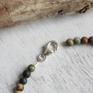 May include: Close-up of a beaded bracelet with a silver clasp. The beads are various earth tones, including green, brown, and tan. The bracelet is on a white wooden surface with a piece of driftwood in the background.