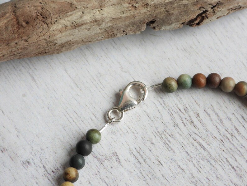 May include: Close-up of a beaded bracelet with a silver clasp. The beads are various earth tones, including green, brown, and tan. The bracelet is on a white wooden surface with a piece of driftwood in the background.