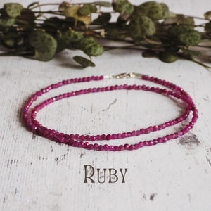 Puede incluir: Un collar de doble cadena hecho de pequeñas cuentas de rubí facetadas. El collar está sobre una superficie de madera blanca. La palabra "Ruby" está escrita debajo del collar.