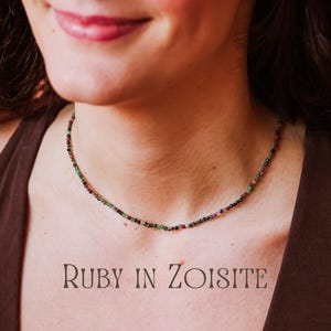 Puede incluir: Primer plano de una mujer que lleva un collar hecho de pequeñas cuentas de colores. Las cuentas son una mezcla de verde, negro y rojo, y el collar está superpuesto en el cuello de la mujer. El texto "RUBY IN ZOISITE" está escrito debajo del collar.