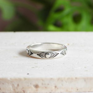 May include: A silver ring with an engraved geometric pattern. The ring features repeating triangular and circular designs around the band. The ring is displayed on a light-colored surface with a blurred green background.