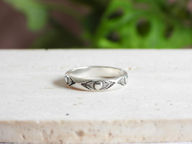 May include: A silver ring with an engraved geometric pattern. The ring features repeating triangular and circular designs around the band. The ring is displayed on a light-colored surface with a blurred green background.