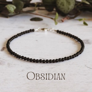 May include: A black obsidian beaded bracelet with a silver clasp. The bracelet is on a white wooden surface. OBSIDIAN is written in black text below the bracelet.