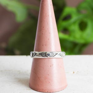 May include: A silver ring with a repeating geometric pattern of diamond and dot shapes. The ring is displayed on a pink cone-shaped stand, with a blurred green plant background. The ring is a band style.