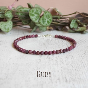 May include: A delicate bracelet composed of small, faceted, deep red ruby beads arranged in a circular pattern. The piece features a silver-toned metal lobster claw clasp closure. The word Ruby is printed in a dark, serif font below the bracelet.