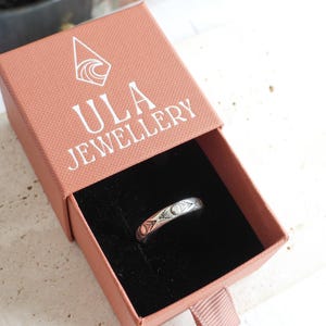 May include: A silver ring with a decorative pattern rests inside an open pink jewelry box. The box has the text "ULA JEWELLERY" printed on the lid. The ring is the focal point, showcasing its intricate design.