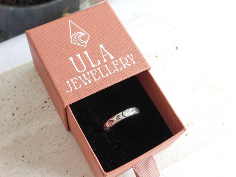 May include: A silver ring with a decorative pattern rests inside an open pink jewelry box. The box has the text "ULA JEWELLERY" printed on the lid. The ring is the focal point, showcasing its intricate design.