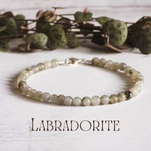 Puede incluir: Un brazalete de plata con cuentas de labradorita grises. El brazalete está sobre una superficie de madera blanca con hojas verdes en el fondo. La palabra "LABRADORITE" está escrita en letras negras debajo del brazalete.