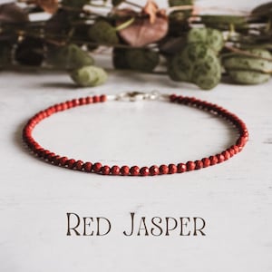 Puede incluir: Un brazalete de cuentas de jaspe rojo con cierre de plata. El brazalete está sobre un fondo blanco. Jaspe rojo.