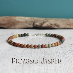 May include: Picasso Jasper beaded bracelet with a silver clasp. The bracelet features small, round beads in shades of brown, orange, green, and gray. The text "Picasso Jasper" is displayed below the bracelet.