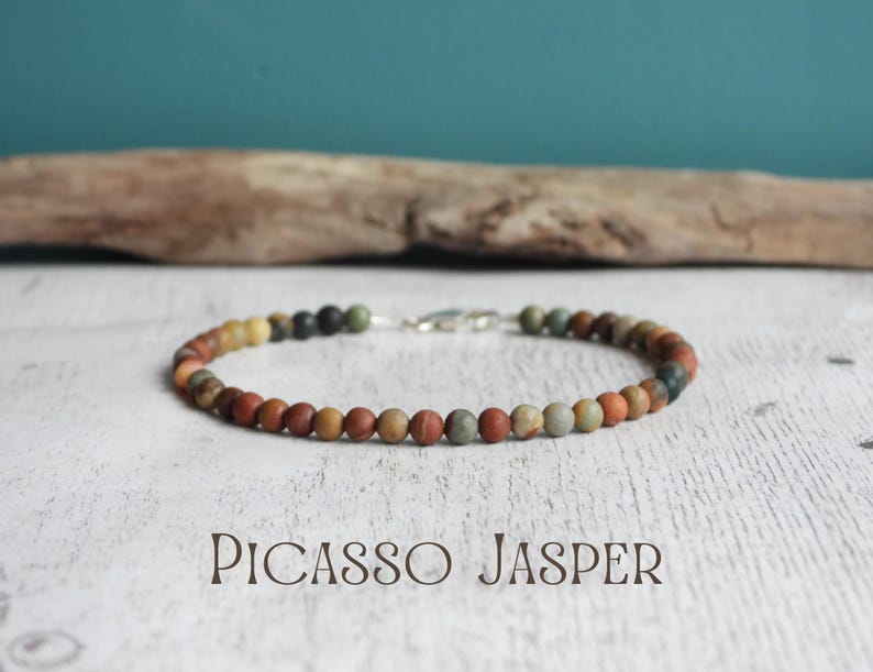 May include: Picasso Jasper beaded bracelet with a silver clasp. The bracelet features small, round beads in shades of brown, orange, green, and gray. The text "Picasso Jasper" is displayed below the bracelet.