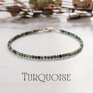 May include: A turquoise beaded bracelet with a silver clasp. The bracelet features small, round beads in shades of green, blue, and brown. The word "TURQUOISE" is printed in a serif font below the bracelet.