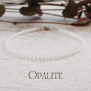 Puede incluir: Un delicado brazalete de opalita con pequeñas cuentas redondas e iridiscentes. El brazalete se exhibe sobre una superficie clara y texturizada. La palabra "OPALITE" está impresa en marrón debajo.