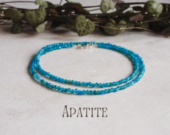 Puede incluir: Un collar de doble cadena hecho de cuentas de apatita azul. El collar está sobre una superficie blanca con follaje verde en el fondo. La palabra "APATITE" está escrita en texto negro debajo del collar.