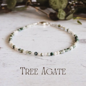 May include: A delicate necklace made with white and green tree agate beads. The necklace is on a white wooden surface. "Tree Agate" is written in black text below the necklace.
