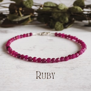 Peut inclure: Un bracelet en perles de rubis rose avec un fermoir argenté. Le bracelet est posé sur une surface en bois blanc. RUBY