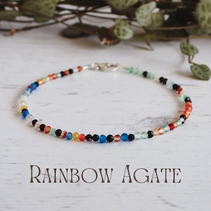 Puede incluir: Un brazalete de cuentas de ágata arcoíris con cuentas negras, blancas, azules, verdes, naranjas y rojas. El brazalete está sobre una superficie de madera blanca.