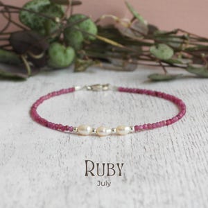 May include: A delicate ruby and pearl bracelet. The bracelet features small, round, deep pink ruby beads strung together, with three oval pearls and a silver clasp. The word "Ruby" and the month "July" are printed below the bracelet.