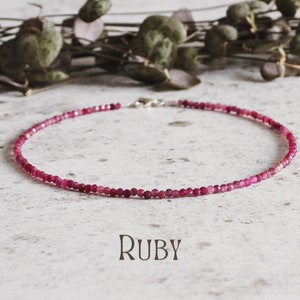 May include: A delicate necklace made with small, round ruby beads. The necklace is on a white background with a leafy branch in the background. The word "RUBY" is written in black below the necklace.