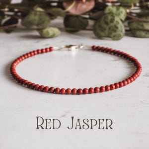 May include: A red jasper beaded bracelet with a silver clasp. The bracelet is on a white surface. "RED JASPER" is written in black text below the bracelet.