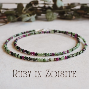 May include: A delicate necklace made with green and pink gemstone beads. The necklace features a mix of small, round beads in shades of green, pink, and black. The text "RUBY IN ZOISITE" is printed below the necklace.