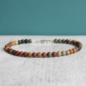 May include: A beaded bracelet featuring small, round beads in a variety of earth tones, including red, brown, yellow, and green. The bracelet is strung on a cord and secured with a silver clasp. The bracelet is approximately 7 inches in length.