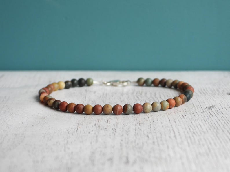May include: A beaded bracelet featuring small, round beads in a variety of earth tones, including red, brown, yellow, and green. The bracelet is strung on a cord and secured with a silver clasp. The bracelet is approximately 7 inches in length.
