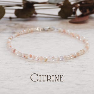 May include: A delicate Citrine bracelet, composed of small, faceted beads in peach and pale yellow hues. The bracelet is strung on a thin cord and fastened with a silver clasp. The word "Citrine" is printed below the bracelet.