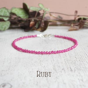 May include: A delicate bracelet composed of small, faceted, vibrant pink ruby beads arranged in a circular pattern. The jewellery piece features a metallic silver-toned clasp mechanism at the top. The word Ruby is printed in a dark font below the item.