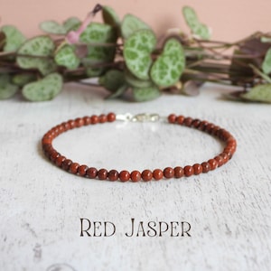 Puede incluir: Una pulsera compuesta por pequeñas cuentas redondas de jaspe rojo oscuro con un cierre de metal plateado. El texto "RED JASPER" está impreso en una fuente serif oscura debajo de la joya, dispuesta en forma circular sobre una superficie blanca.