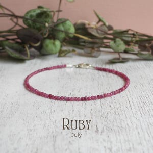 May include: A fine ruby bracelet with a silver clasp. The bracelet is composed of small, round, deep pink gemstones. The word "RUBY" is printed in a serif font below the bracelet, with "July" underneath.