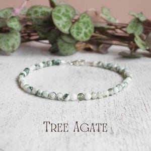May include: A delicate bracelet composed of small, round tree agate beads featuring a mottled pattern of white and forest green. The bracelet is finished with a silver-toned metal lobster clasp and is labelled with the text TREE AGATE.