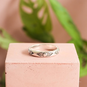 May include: A silver ring with an engraved pattern of triangles and half-moon shapes. The ring is displayed on a small, square, pink block, with a blurred green plant in the background.