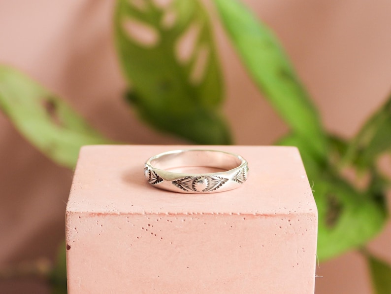 May include: A silver ring with an engraved pattern of triangles and half-moon shapes. The ring is displayed on a small, square, pink block, with a blurred green plant in the background.
