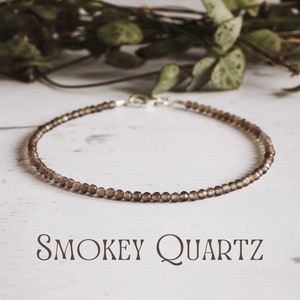 May include: A delicate bracelet made with smoky quartz beads and a silver clasp. The bracelet is on a white wooden surface.