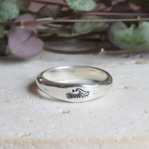 May include: A silver ring with a wave design. The ring is smooth and polished, with a small, engraved wave and star pattern. The ring is set against a neutral background with green foliage.